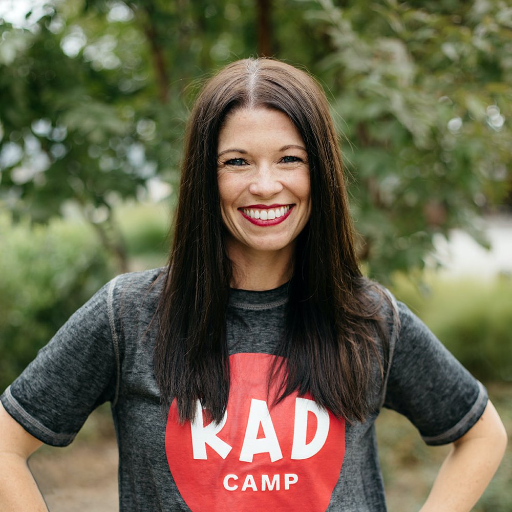 A white woman with dark brown hair and red lipstick smiling. She is wearing a grey t-shirt with the RAD Camp logo in a pink circle.