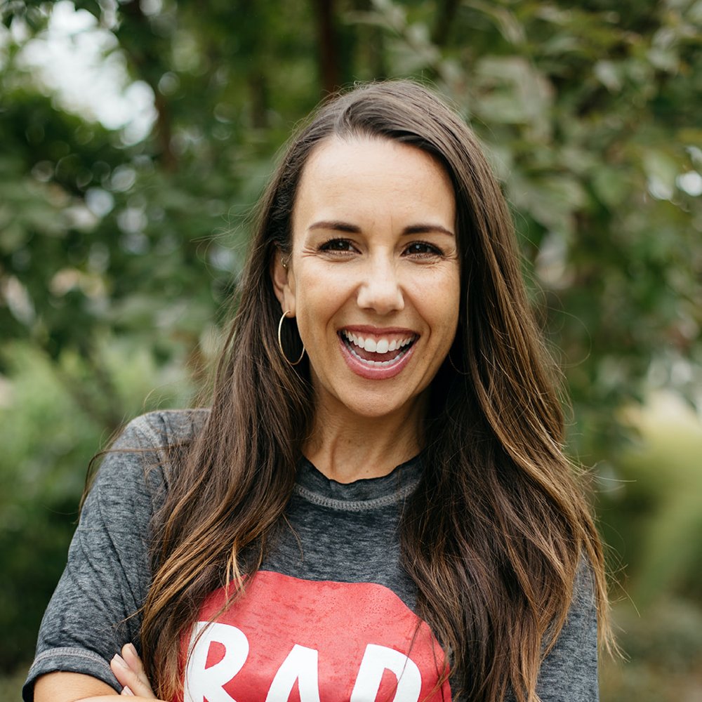 A white woman with dark brown hair has her arms across her chest and is smiling. She is wearing a grey t-shirt with the RAD Camp logo in a pink circle.