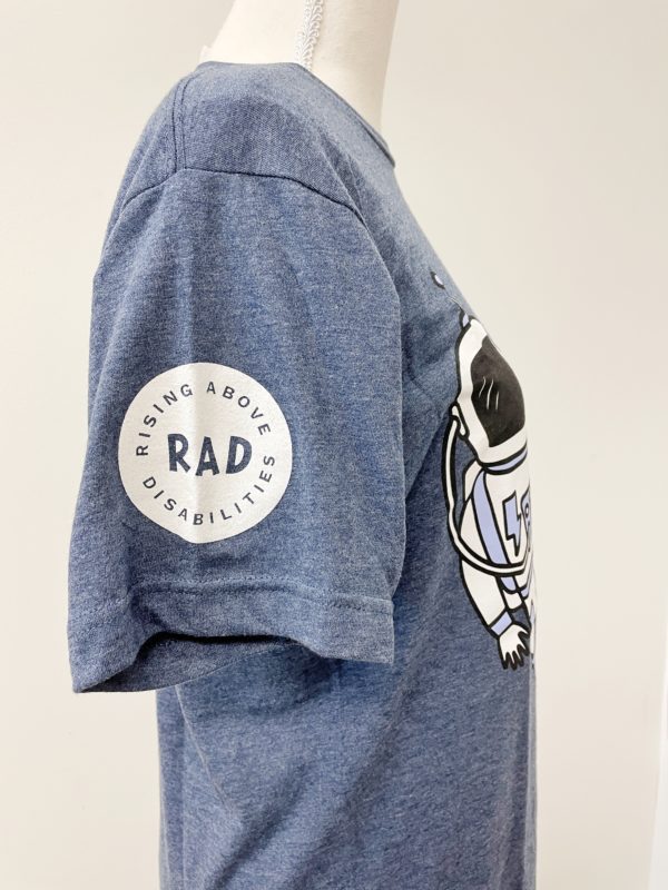 Side of heather gray blue shirt. Sleeve has a white circle. The words RAD is written in the middle and the words Rising Above Disabilities is written in a circle around RAD.