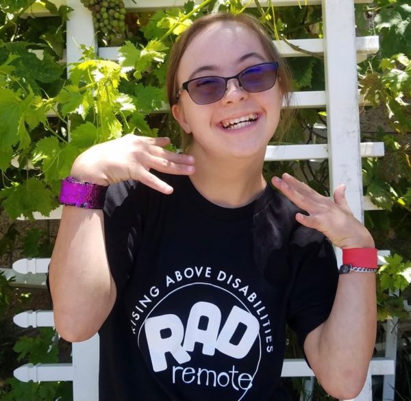 A smiling girl with tinted glasses poses with her hands near her face. She is wearing a black t-shirt with white letters which reads RAD Remote in a circle. Rising Above Disabilities is along the top of the circle