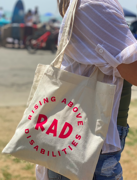Image of a woman below the head wearing a reusable bag on her shoulder. The word RAD is in the middle with the pink words Rising Above Disabilities around it in a circle.