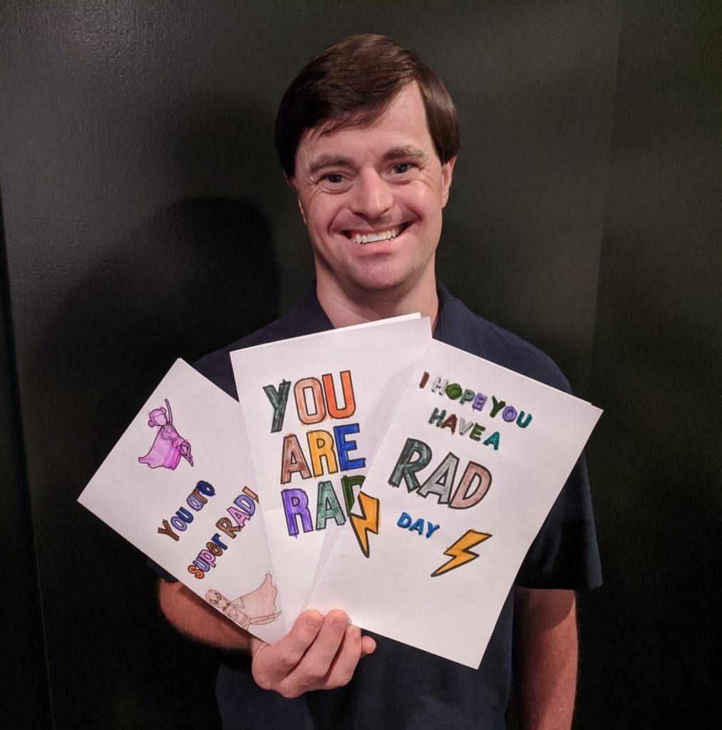 A man smiling and holding up 3 colored in cards that have RAD messages on them