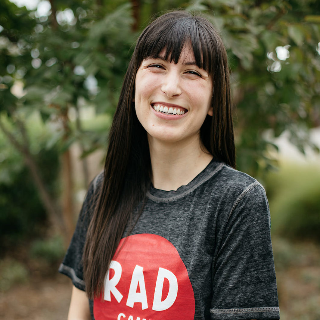An asian-hispanic woman with dark brown hair smiling. She is wearing a grey t-shirt with the RAD Camp logo in a pink circle.